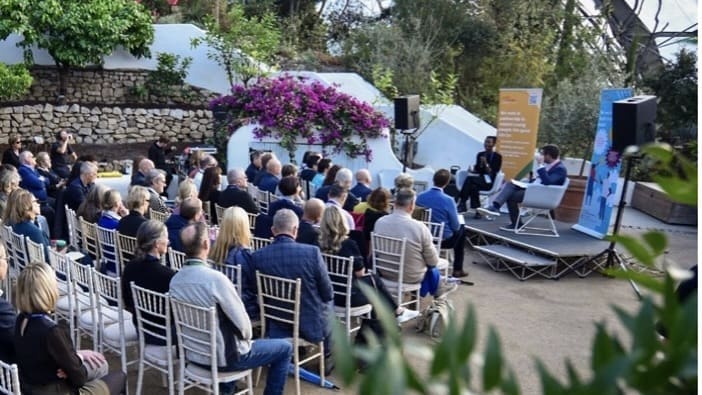 People sat on chairs in front of a stage in a greenhouse